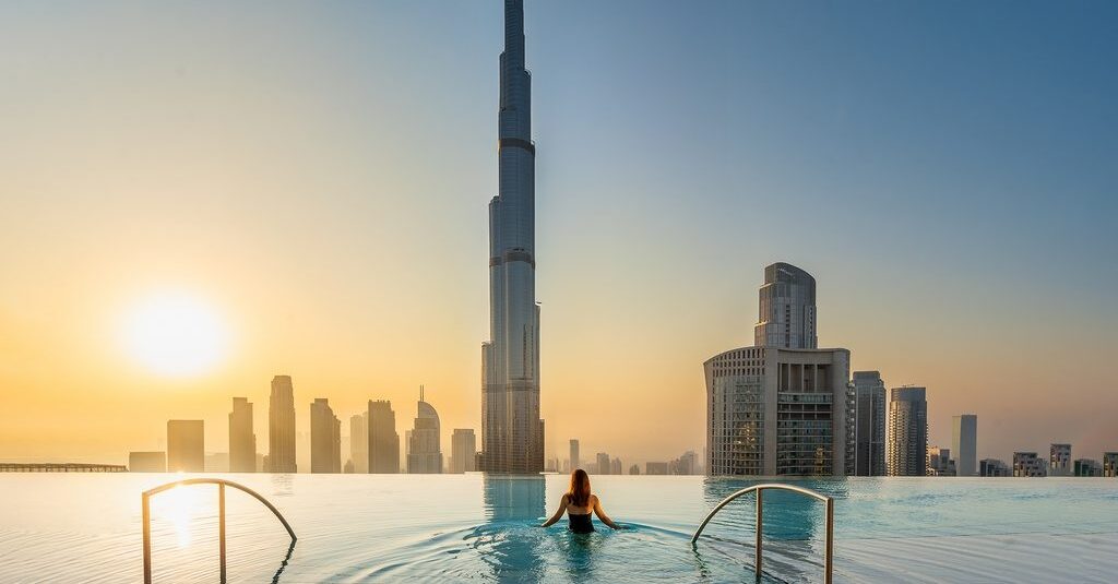 Foto panorâmica de um luxuoso beach club em Dubai ao pôr do sol, com piscina de borda infinita voltada para o mar cristalino e o skyline moderno da cidade ao fundo. Espreguiçadeiras elegantes, palmeiras iluminadas e atmosfera sofisticada transmitem exclusividade, turismo de luxo e experiência premium nos Emirados Árabes Unidos.