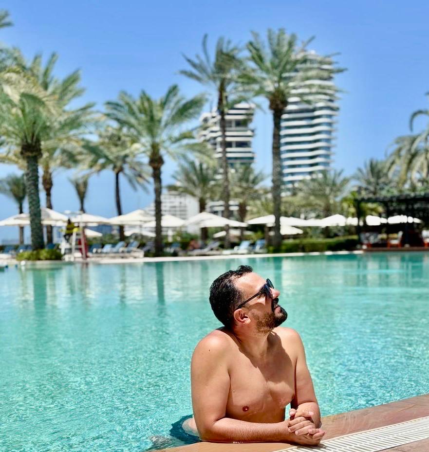 Foto panorâmica de um luxuoso beach club em Dubai ao pôr do sol, com piscina de borda infinita voltada para o mar cristalino e o skyline moderno da cidade ao fundo. Espreguiçadeiras elegantes, palmeiras iluminadas e atmosfera sofisticada transmitem exclusividade, turismo de luxo e experiência premium nos Emirados Árabes Unidos.