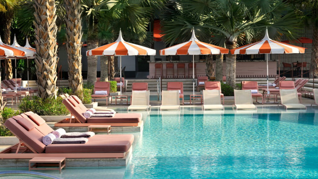 Foto panorâmica de um luxuoso beach club em Dubai ao pôr do sol, com piscina de borda infinita voltada para o mar cristalino e o skyline moderno da cidade ao fundo. Espreguiçadeiras elegantes, palmeiras iluminadas e atmosfera sofisticada transmitem exclusividade, turismo de luxo e experiência premium nos Emirados Árabes Unidos.