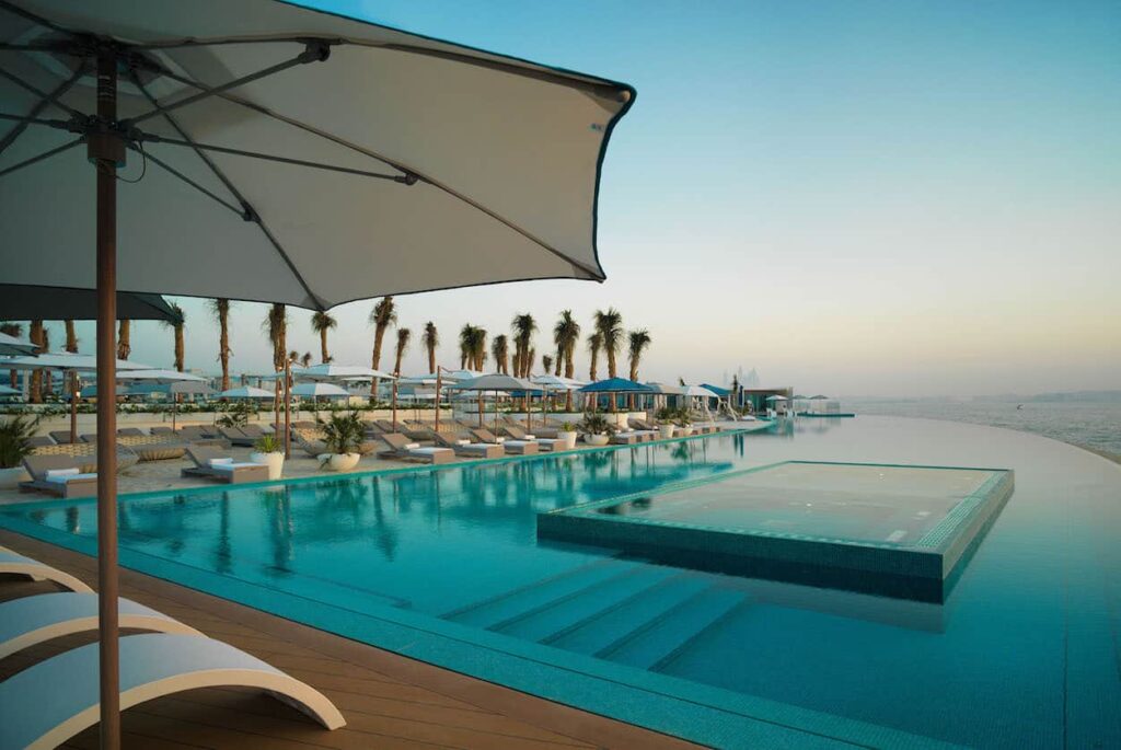 Foto panorâmica de um luxuoso beach club em Dubai ao pôr do sol, com piscina de borda infinita voltada para o mar cristalino e o skyline moderno da cidade ao fundo. Espreguiçadeiras elegantes, palmeiras iluminadas e atmosfera sofisticada transmitem exclusividade, turismo de luxo e experiência premium nos Emirados Árabes Unidos.