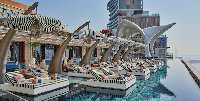 Foto panorâmica de um luxuoso beach club em Dubai ao pôr do sol, com piscina de borda infinita voltada para o mar cristalino e o skyline moderno da cidade ao fundo. Espreguiçadeiras elegantes, palmeiras iluminadas e atmosfera sofisticada transmitem exclusividade, turismo de luxo e experiência premium nos Emirados Árabes Unidos.