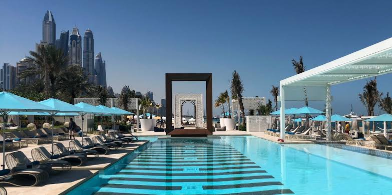Foto panorâmica de um luxuoso beach club em Dubai ao pôr do sol, com piscina de borda infinita voltada para o mar cristalino e o skyline moderno da cidade ao fundo. Espreguiçadeiras elegantes, palmeiras iluminadas e atmosfera sofisticada transmitem exclusividade, turismo de luxo e experiência premium nos Emirados Árabes Unidos.