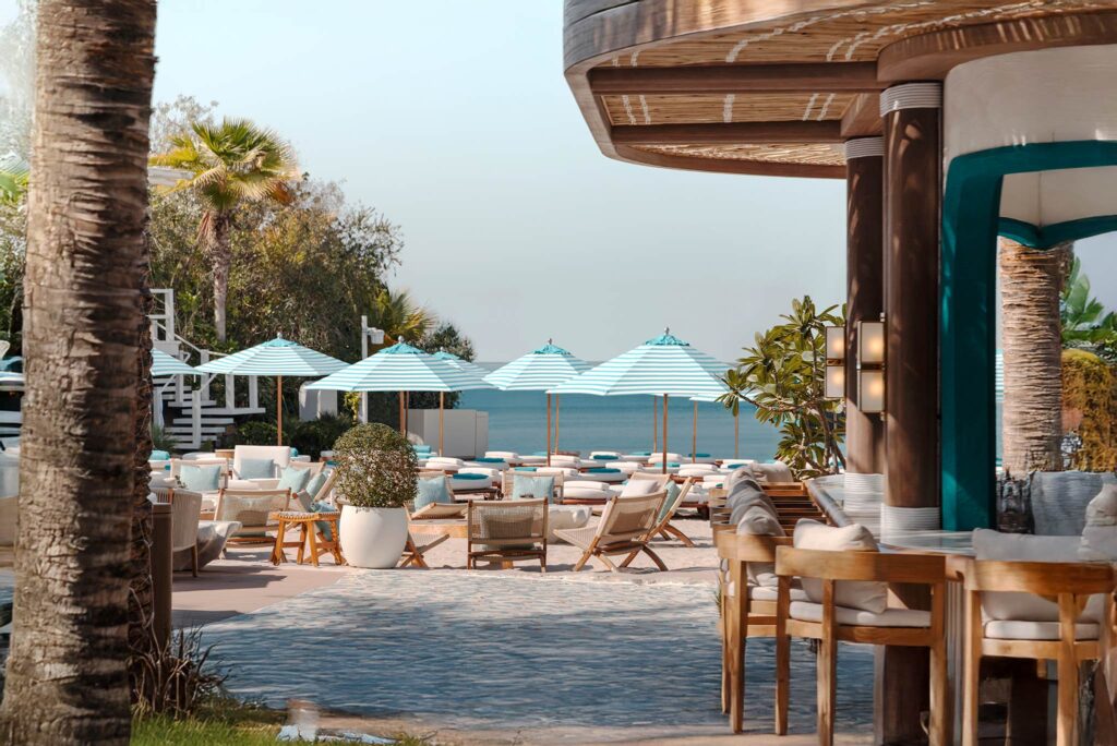 Foto panorâmica de um luxuoso beach club em Dubai ao pôr do sol, com piscina de borda infinita voltada para o mar cristalino e o skyline moderno da cidade ao fundo. Espreguiçadeiras elegantes, palmeiras iluminadas e atmosfera sofisticada transmitem exclusividade, turismo de luxo e experiência premium nos Emirados Árabes Unidos.