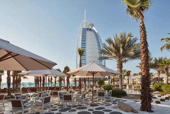 Foto panorâmica de um luxuoso beach club em Dubai ao pôr do sol, com piscina de borda infinita voltada para o mar cristalino e o skyline moderno da cidade ao fundo. Espreguiçadeiras elegantes, palmeiras iluminadas e atmosfera sofisticada transmitem exclusividade, turismo de luxo e experiência premium nos Emirados Árabes Unidos.