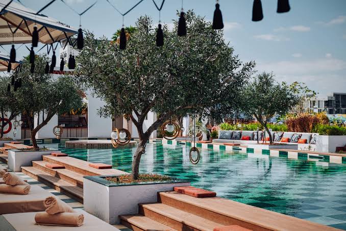 Foto panorâmica de um luxuoso beach club em Dubai ao pôr do sol, com piscina de borda infinita voltada para o mar cristalino e o skyline moderno da cidade ao fundo. Espreguiçadeiras elegantes, palmeiras iluminadas e atmosfera sofisticada transmitem exclusividade, turismo de luxo e experiência premium nos Emirados Árabes Unidos.