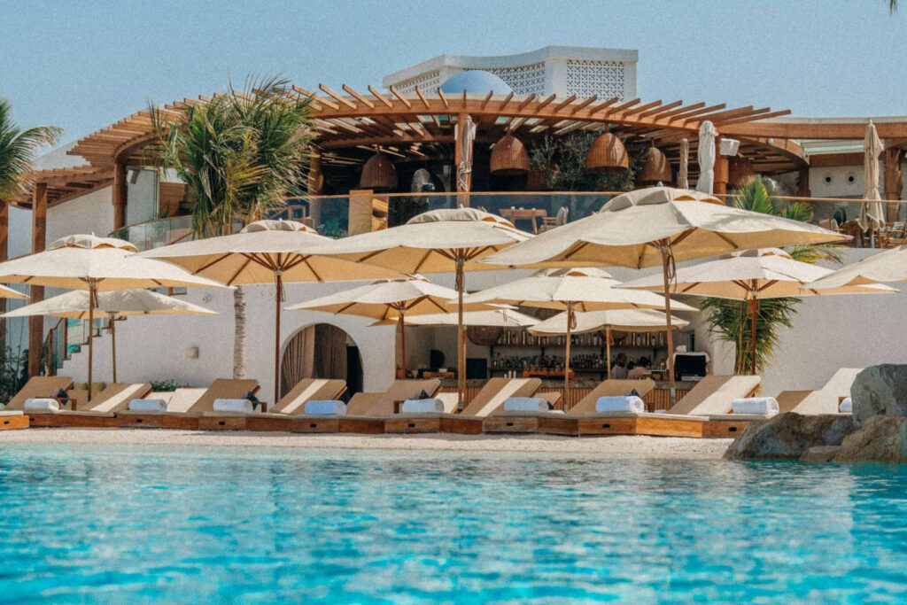 Foto panorâmica de um luxuoso beach club em Dubai ao pôr do sol, com piscina de borda infinita voltada para o mar cristalino e o skyline moderno da cidade ao fundo. Espreguiçadeiras elegantes, palmeiras iluminadas e atmosfera sofisticada transmitem exclusividade, turismo de luxo e experiência premium nos Emirados Árabes Unidos.