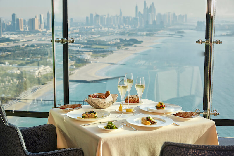 Foto panorâmica de um restaurante luxuoso em Dubai, com vista para o skyline moderno da cidade ao fundo. Ambiente sofisticado com mesas elegantes, decoração refinada e atmosfera exclusiva que transmite uma experiência gastronômica premium e turismo de luxo nos Emirados Árabes Unidos.