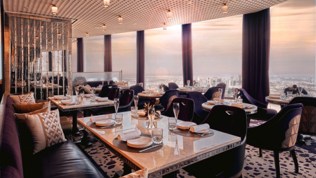 Foto panorâmica de um restaurante luxuoso em Dubai, com vista para o skyline moderno da cidade ao fundo. Ambiente sofisticado com mesas elegantes, decoração refinada e atmosfera exclusiva que transmite uma experiência gastronômica premium e turismo de luxo nos Emirados Árabes Unidos.