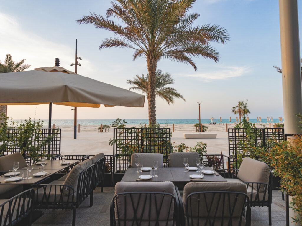 Foto panorâmica de um restaurante italiano luxuoso em Dubai, com salão elegante, mesas brancas impecavelmente montadas, iluminação suave, serviço refinado e ambiente exclusivo no coração financeiro da cidade. Atmosfera clássica, sofisticada e premium, ideal para turismo de luxo, jantares especiais e encontros de negócios nos Emirados Árabes Unidos.