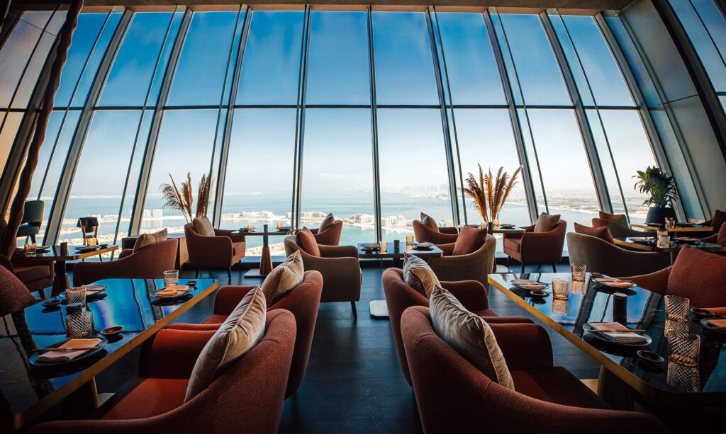 Foto panorâmica de um restaurante luxuoso em Dubai, com vista para o skyline moderno da cidade ao fundo. Ambiente sofisticado com mesas elegantes, decoração refinada e atmosfera exclusiva que transmite uma experiência gastronômica premium e turismo de luxo nos Emirados Árabes Unidos.