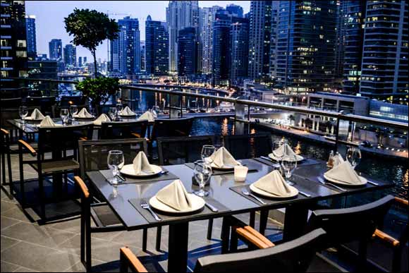 Foto panorâmica de um restaurante árabe luxuoso em Dubai Marina, com mesas elegantes, decoração refinada, pratos libaneses servidos à mesa, iluminação acolhedora & vista para os prédios modernos & o canal da Dubai Marina ao fundo. Ambiente sofisticado que transmite gastronomia árabe premium, turismo de luxo & experiência cultural nos Emirados Árabes Unidos.