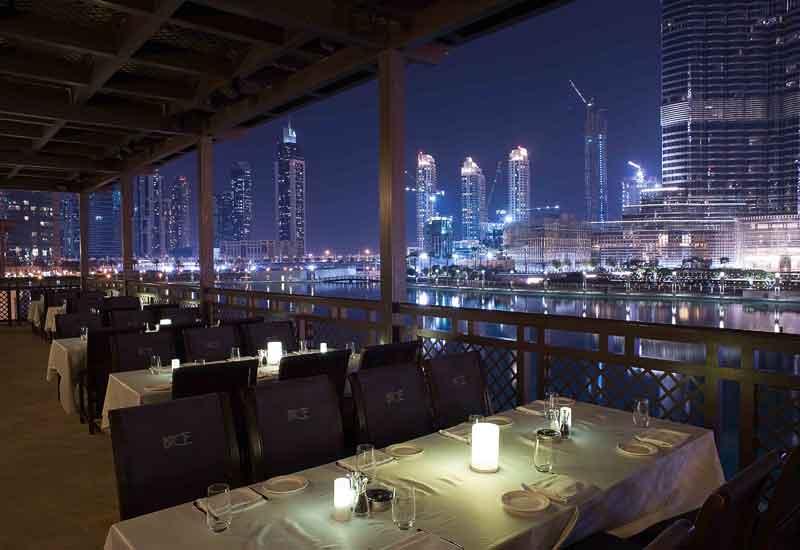 Foto panorâmica de restaurante italiano elegante em Dubai, com frutos do mar frescos, mesas sofisticadas, vista para o Burj Khalifa e para o espetáculo das águas de Dubai ao fundo. Ambiente luxuoso e romântico.