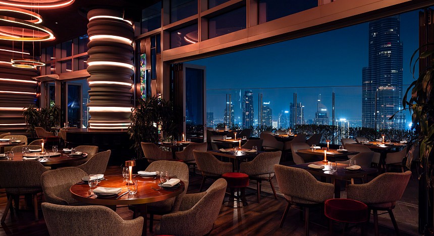 Foto panorâmica de um restaurante luxuoso em Dubai, com vista para o skyline moderno da cidade ao fundo. Ambiente sofisticado com mesas elegantes, decoração refinada e atmosfera exclusiva que transmite uma experiência gastronômica premium e turismo de luxo nos Emirados Árabes Unidos.