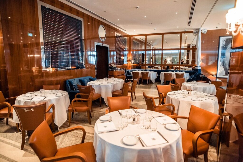 Foto panorâmica de um restaurante italiano luxuoso em Dubai, com salão elegante, mesas brancas impecavelmente montadas, iluminação suave, serviço refinado e ambiente exclusivo no coração financeiro da cidade. Atmosfera clássica, sofisticada e premium, ideal para turismo de luxo, jantares especiais e encontros de negócios nos Emirados Árabes Unidos.