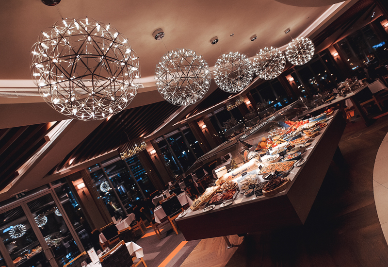 Foto panorâmica de um restaurante luxuoso em Dubai, com vista para o skyline moderno da cidade ao fundo. Ambiente sofisticado com mesas elegantes, decoração refinada e atmosfera exclusiva que transmite uma experiência gastronômica premium e turismo de luxo nos Emirados Árabes Unidos.