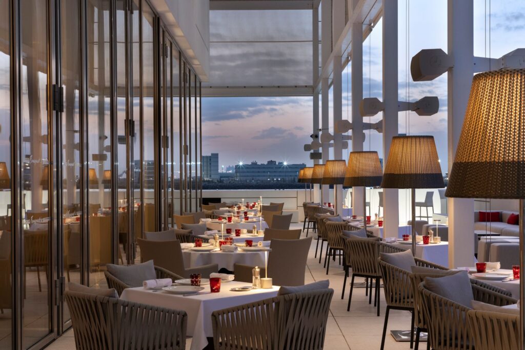 Foto panorâmica de um restaurante italiano luxuoso em Dubai, com salão elegante, mesas brancas impecavelmente montadas, iluminação suave, serviço refinado e ambiente exclusivo no coração financeiro da cidade. Atmosfera clássica, sofisticada e premium, ideal para turismo de luxo, jantares especiais e encontros de negócios nos Emirados Árabes Unidos.