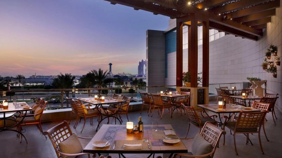Foto panorâmica de um restaurante italiano luxuoso em Dubai, com salão elegante, mesas brancas impecavelmente montadas, iluminação suave, serviço refinado e ambiente exclusivo no coração financeiro da cidade. Atmosfera clássica, sofisticada e premium, ideal para turismo de luxo, jantares especiais e encontros de negócios nos Emirados Árabes Unidos.